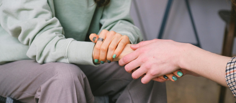 Mão de mulher agarra mão de outra mulher com unhas pintadas de verde, camisola verde-clara e calças cinzas enquanto fala da pensão de sobrevivência.  