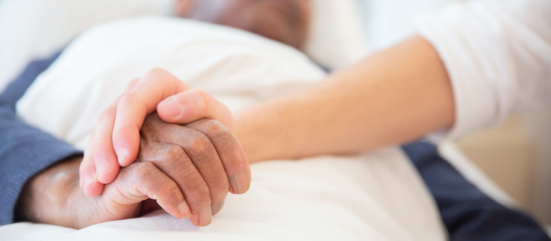 Mão jovem segura na mão de idoso deitado na cama que recebe complemento por dependência.