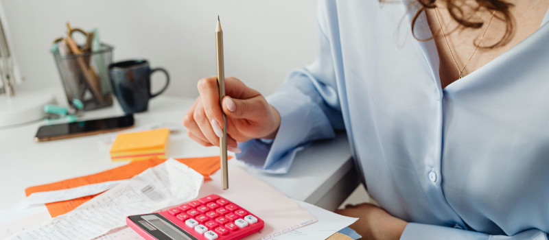 Tronco de jovem mulher de camisa azul, segurando um lápis em cima de calculadora vermelha a pensar como funciona o IRS Jovem.