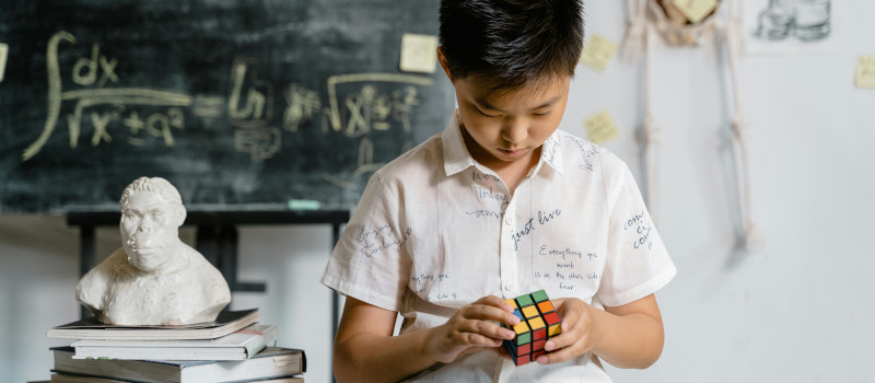 Menino de camisa branca a mexer em cubo de rubik enquanto usa um dos tipos de raciocínio, com um quadro de escola preto atrás, com uma fórmula matemática escrita a giz e uma escultura branca ao lado em cima de uma pilha de livros.